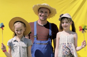 Ein Mann mit Strohhut und Latzhose, rechts und links neben ihm zwei Mädchen mit bunten Plastikwindrädchen